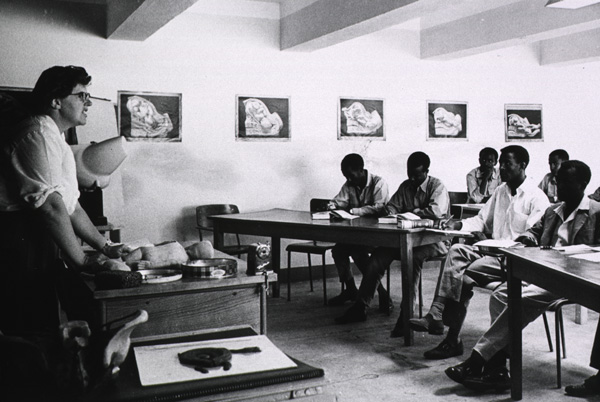 A woman instructor stands facing several men with books seated by desks in a classroom. 