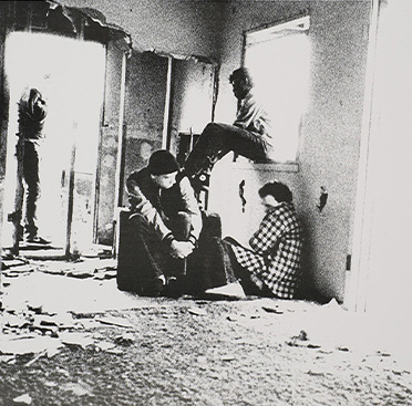 Black and white photograph of run-down front room of a house with three people huddled together, with a fourth person walking out the front door.