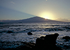 Half of the brightly-shining sun is obscured by a rock formation, but the rest of it can be seen in the sky. Frothy surf bubbles over the rocky beach.