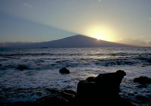 Half of the brightly-shining sun is obscured by a rock formation, but the rest of it can be seen in the sky. Frothy surf bubbles over the rocky beach.