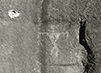 In this black and white photograph, a bullet hole is seen right above an ancient drawing of a person on a rock wall.