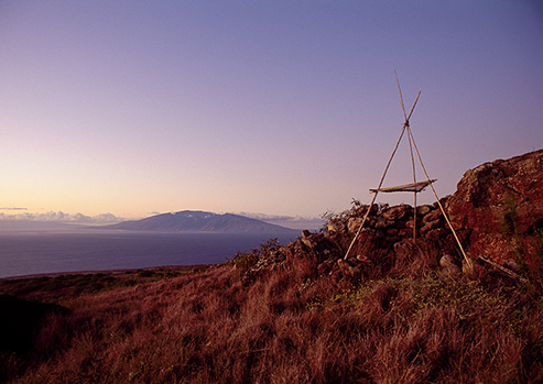 A wooden structure is pictured amidst a rust-colored landscape of scrubby brush and rockiness.