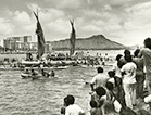 Black-and-white photograph of Hōkūle‘a voyaging canoe returning from its first voyage to Tahiti in 1976. Spectators watch the Hōkūle‘a coming into harbor as other canoes surround it.