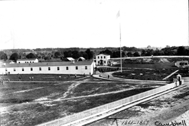 Black and white photograph of a group of wooden buildings with trees and tents in the background and a flagpole in front of one building. Courtesy The Historical Society of Washington, D.C.