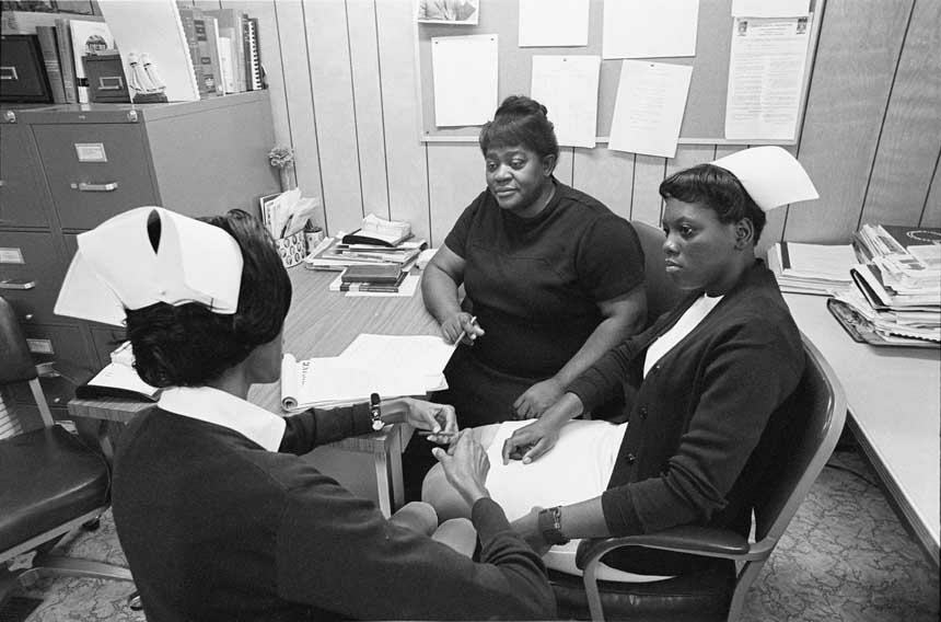 Three African American women sit around a desk; two are wearing nursing caps.