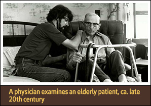 Seated White man providing care to a seated White man in a chair with a walker in front of him.