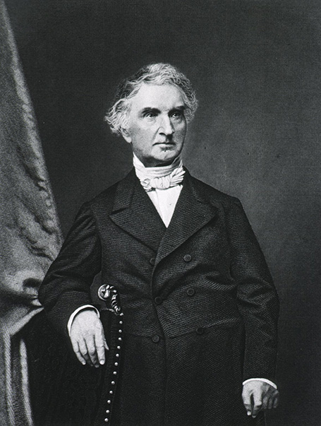 Half-length portrait of a man standing with the right arm over back of a chair.