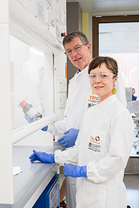 A white man and woman in lab coats face a whiteboard while looking into the camera.  