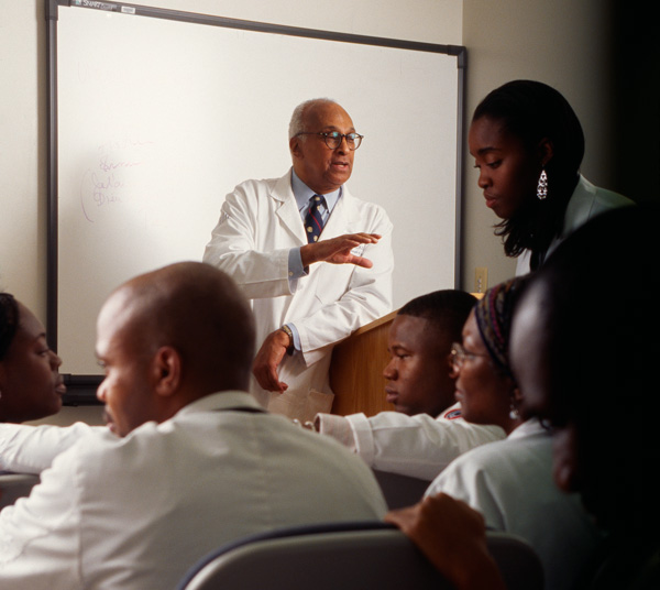 A man leans against a lectern speaking to people seated in a classroom.