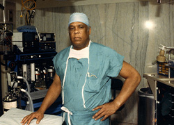 Man in green surgical scrubs and head covering standing in an operating room.