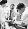 Two White men tend to a mannequin as two White women stand by, one of whom is instructing.