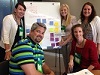 4 White women and 1 White man, stand and sit by a board with post it notes attached.