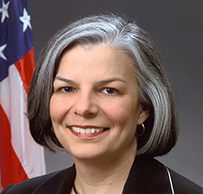 Dr. Julie Louise Gerberding, a White female in a dark suit posing in front of the American flag.