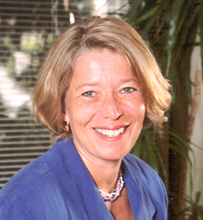 Dr. Linda Rosenstock, a White female in a blue dress posing in an office with a table and palm tree.