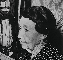 Dr. Lillian Heath Nelson, an elderly female in a polka dot dress holding an object at a desk with books and papers.