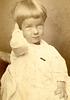 A young girl in dress, seated with right hand behind head, looks slightly to the right of viewer.