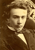 Young man in a suit and bow tie, seated in ornate chair looking down and to the right of the viewer.