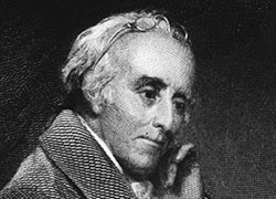 Formal portrait of a white man seated at a desk with an open book; glasses on forehead; thoughtful pose.